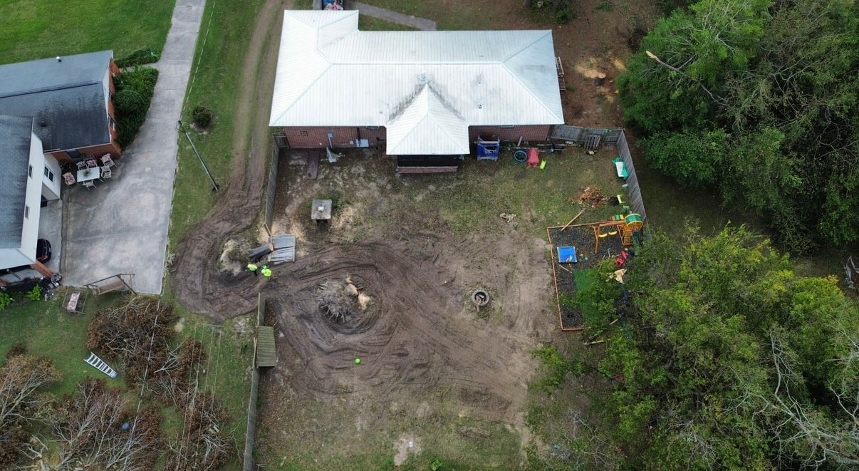 Overhead view of a house with a metal roof and muddy backyard, with scattered debris and two adjacent houses.
