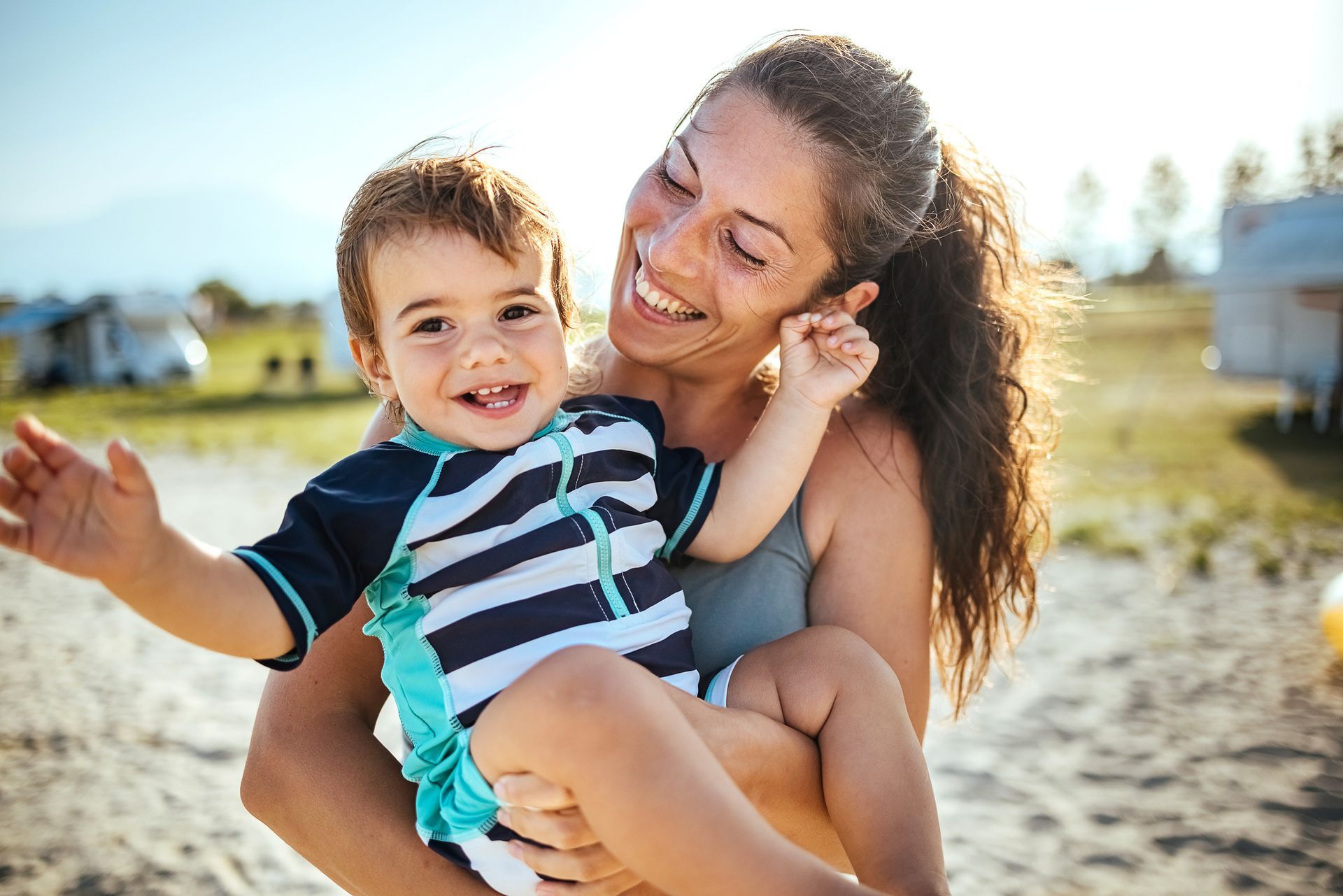 A mother is holding her child and they are both smiling. They are outside on a beach.
