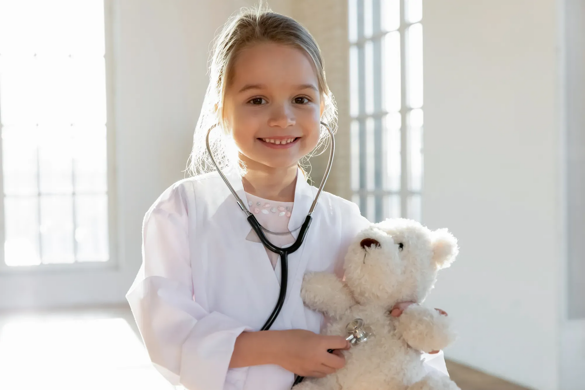 A little girl dressed as a doctor is holding a teddy bear and listening to it with a stethoscope.