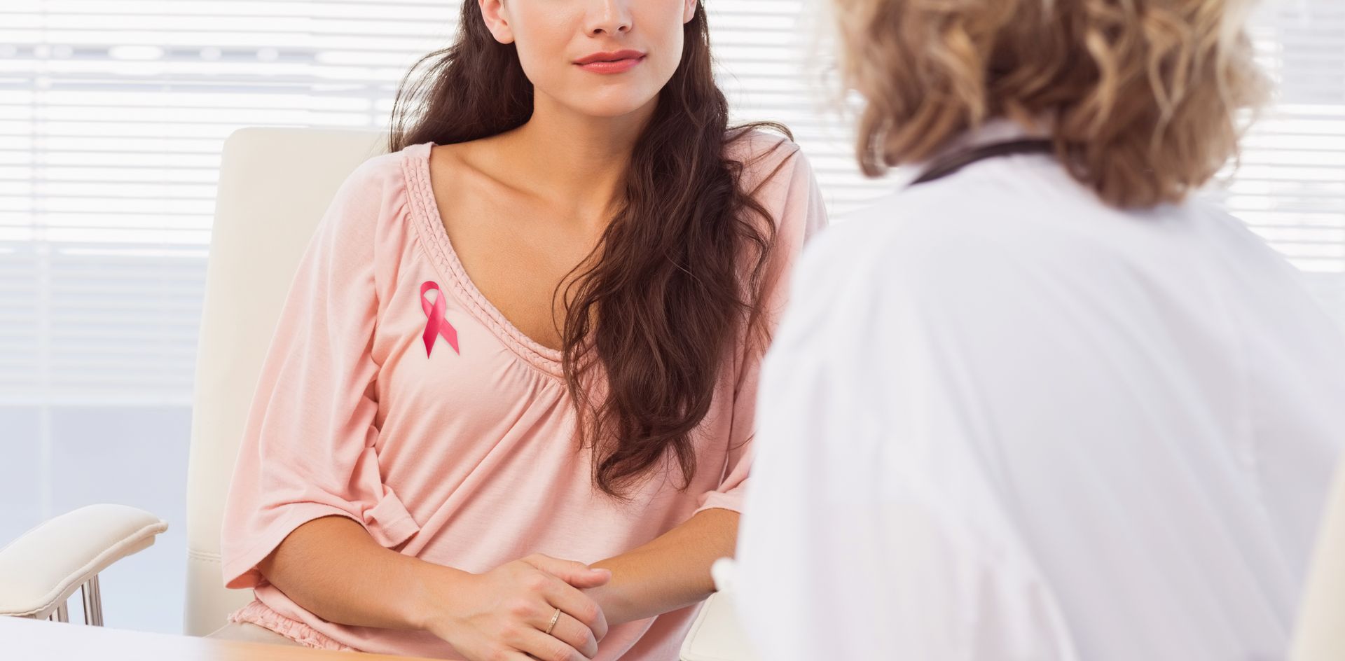 A woman with a pink ribbon on her shirt is talking to a doctor.