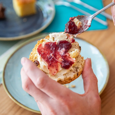 fresh fruit scone with clotted on the base, topped with jam. The scone is being held by a hand.