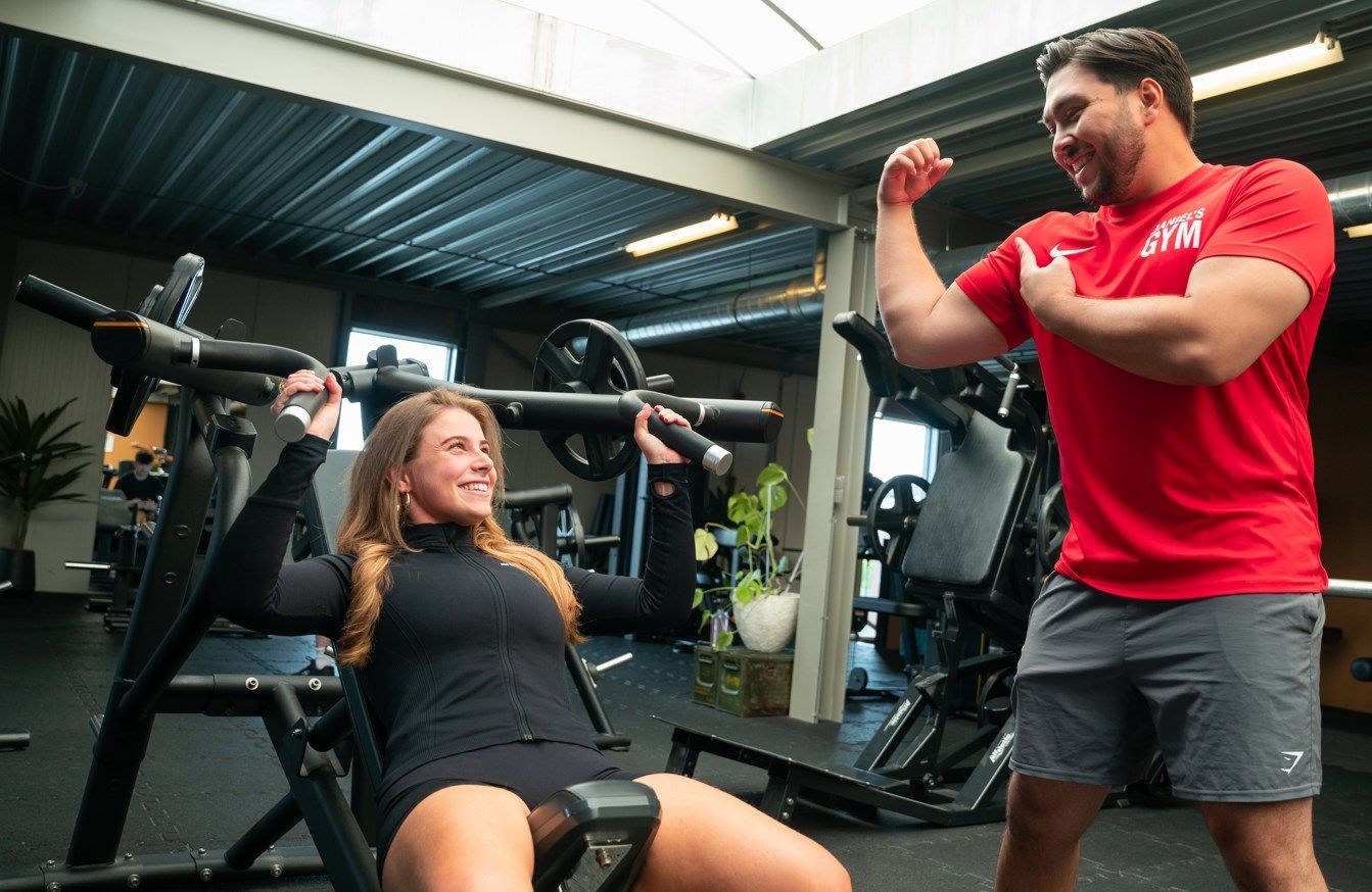 Vrouw die fitnessapparatuur gebruikt en glimlacht, terwijl een man die zijn biceps aanspant toekijkt