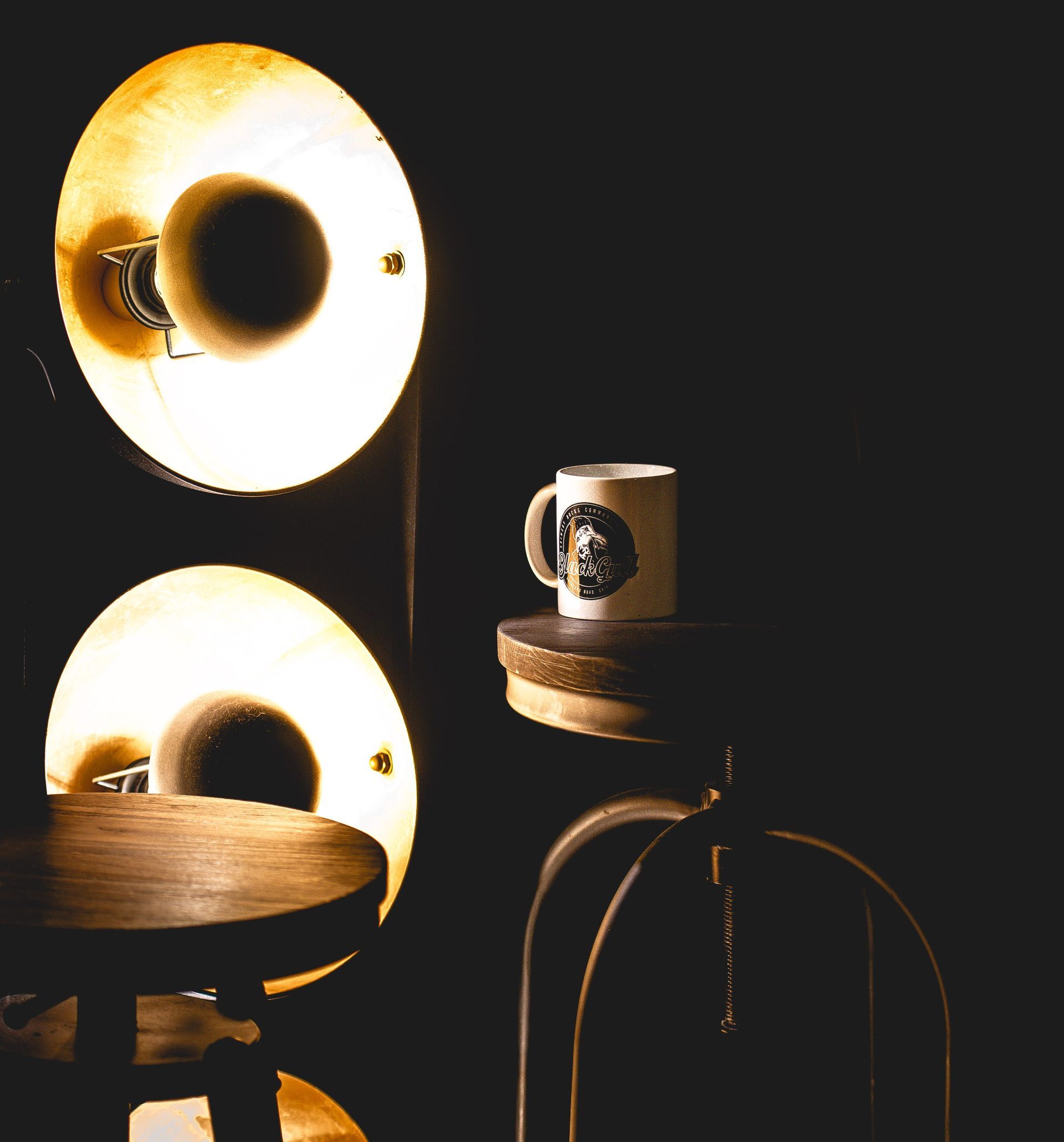 Two bright lamps illuminating a coffee mug on a small shelf near a stool. Dark background.