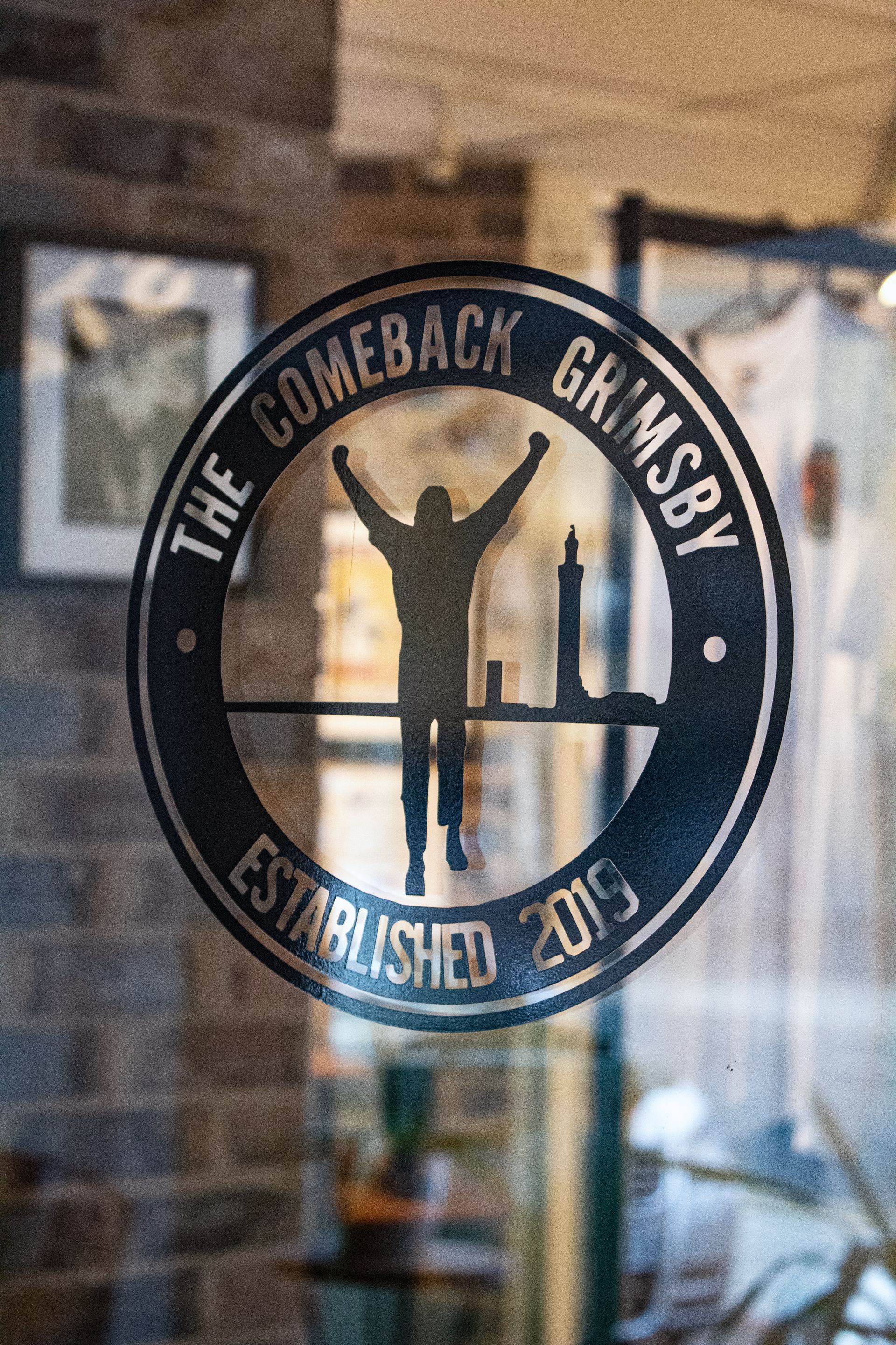Circular sign for "The Comeback Grimsby," with a silhouetted person celebrating, and a lighthouse. Established 2019.