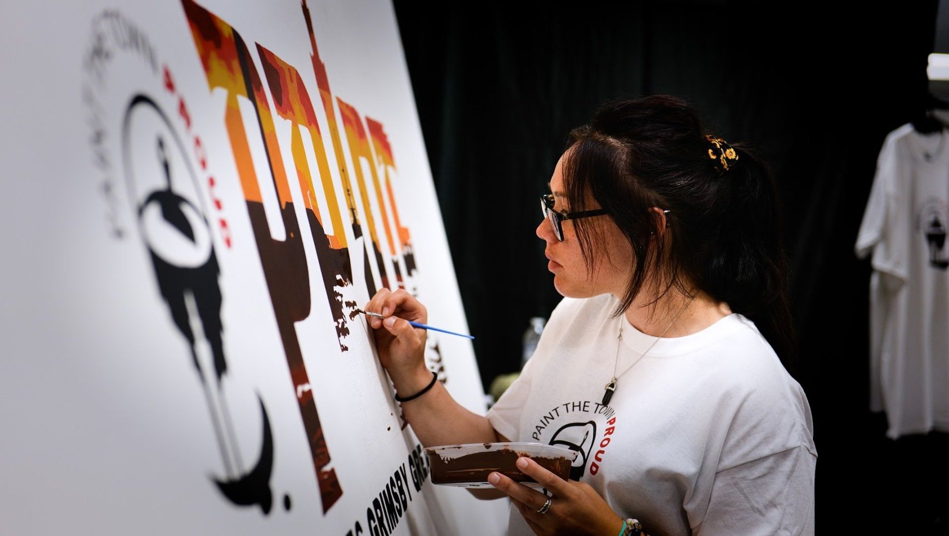 Woman painting on a large white canvas, brown paint, wearing glasses and white t-shirt.