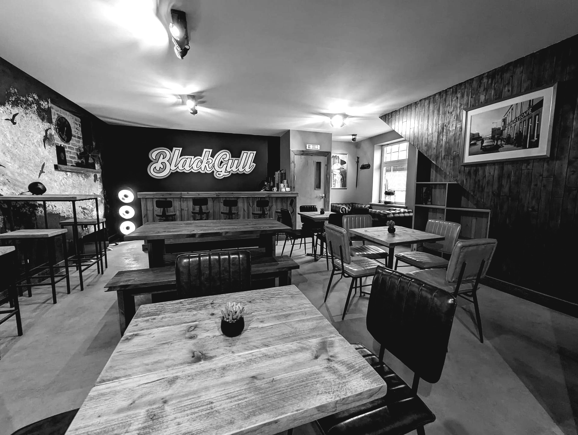 Black and white photo of a dimly lit bar interior, with wooden tables, bar, chairs, and wall art.