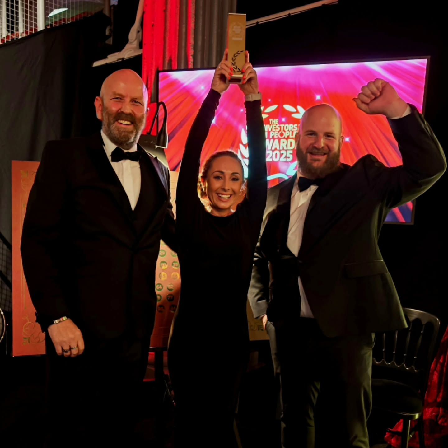 Three people celebrating an award; woman holding it up, two men on either side. Stage backdrop.