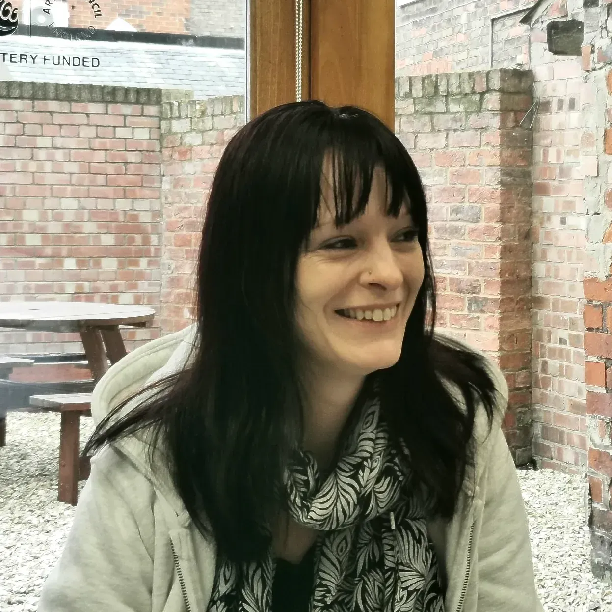 Woman smiling outdoors, wearing a light gray hoodie and patterned scarf, in front of brick walls and a picnic table.
