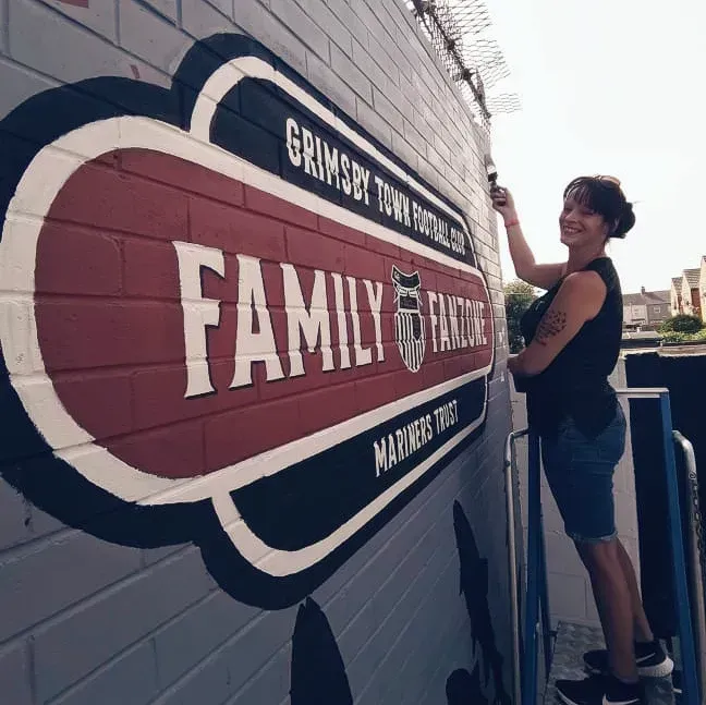 Woman paints a Grimsby Town Football Club mural on a brick wall, smiling.