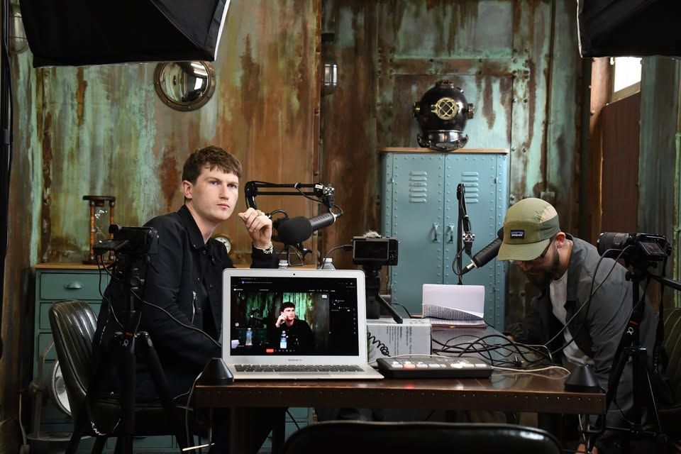 Film crew in a rustic studio, with a laptop, camera gear, and two people working at a table.