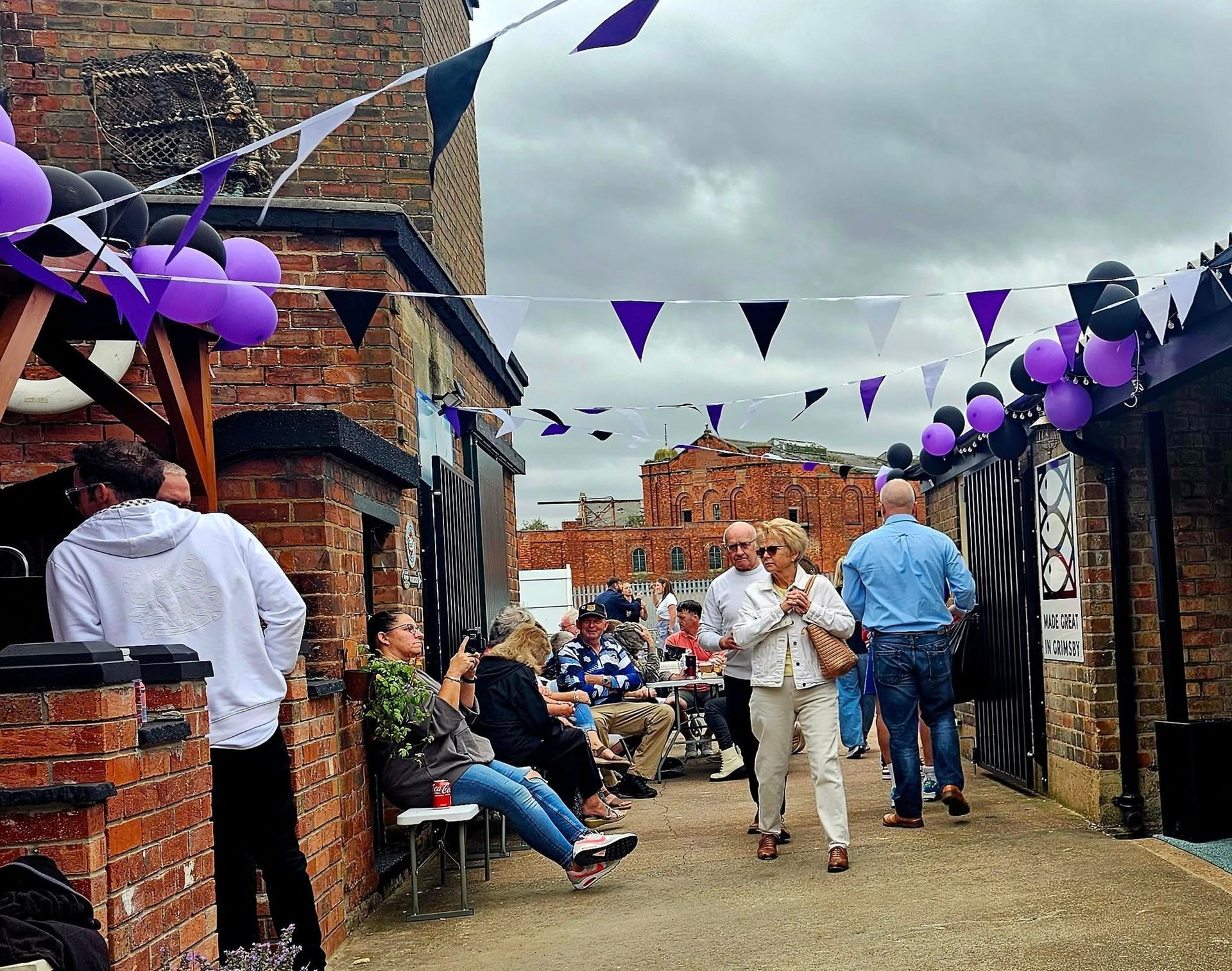 People at an outdoor event, brick buildings, purple and black decorations, cloudy sky.
