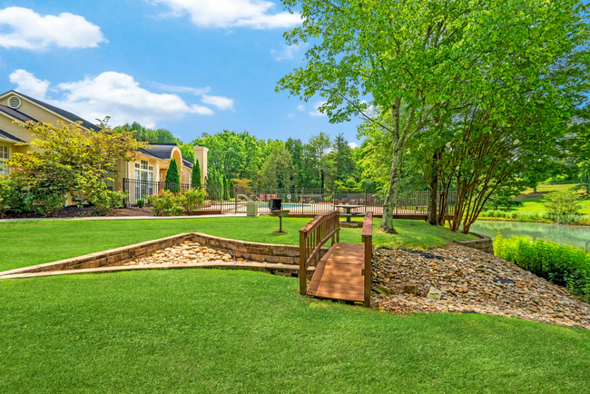A wooden bridge over a river in a park with a house in the background.