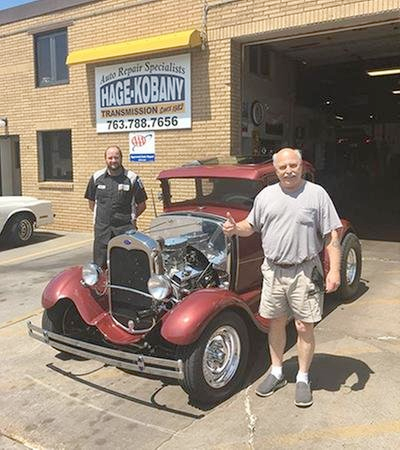 Two men are standing next to a red car in front of a garage.