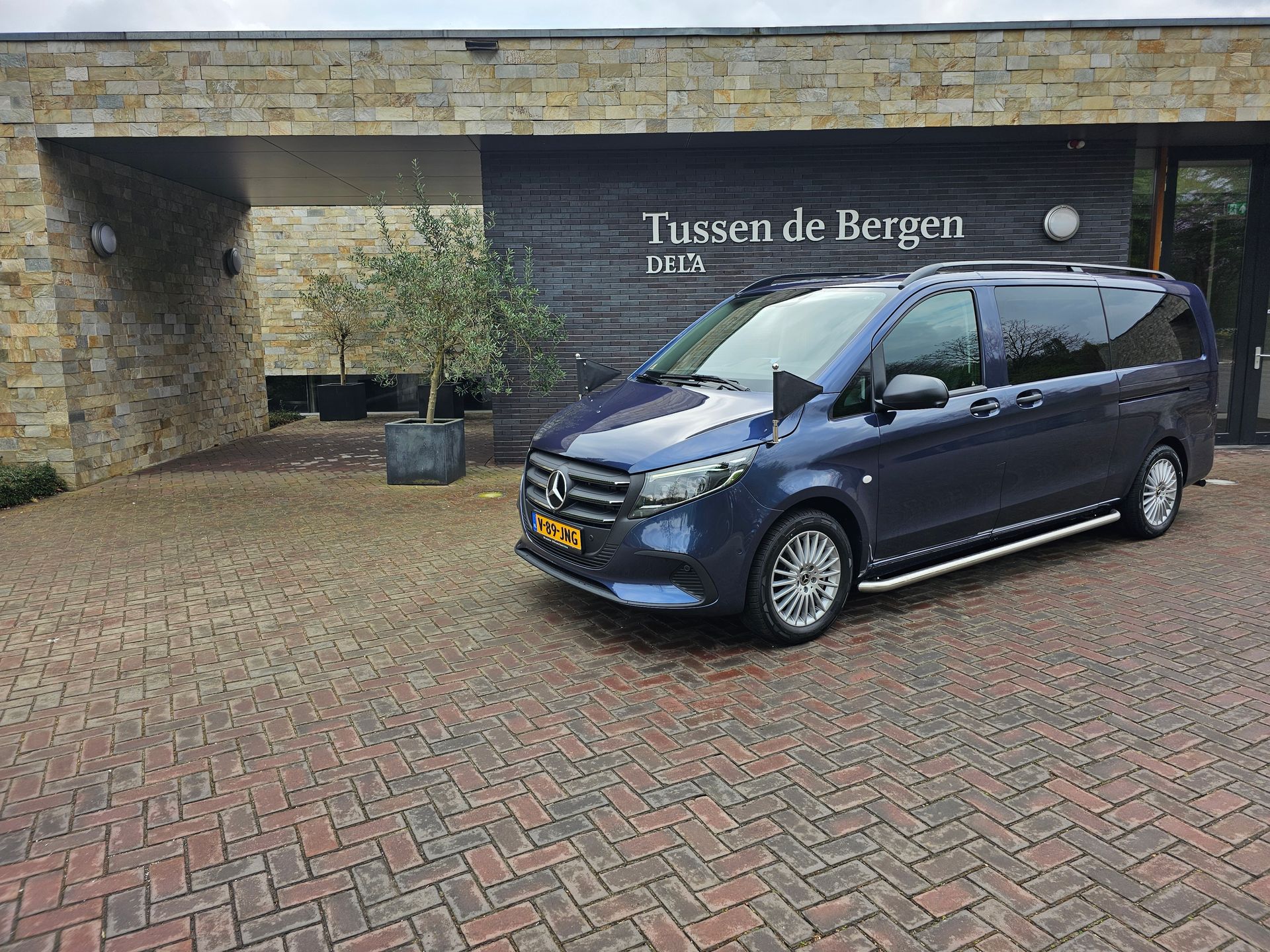 Ciemnoniebieski van Mercedes-Benz zaparkowany na wyasfaltowanym podjeździe przed nowoczesnym budynkiem o nazwie „Tussen de Bergen”.