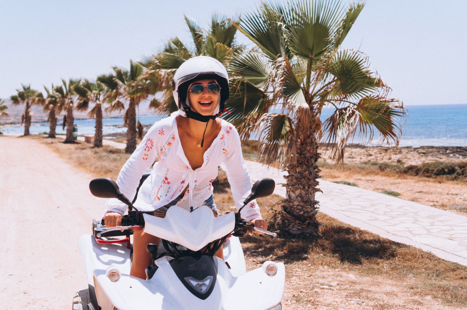 Woman on white scooter, wearing helmet and sunglasses, smiles near palm trees and ocean.