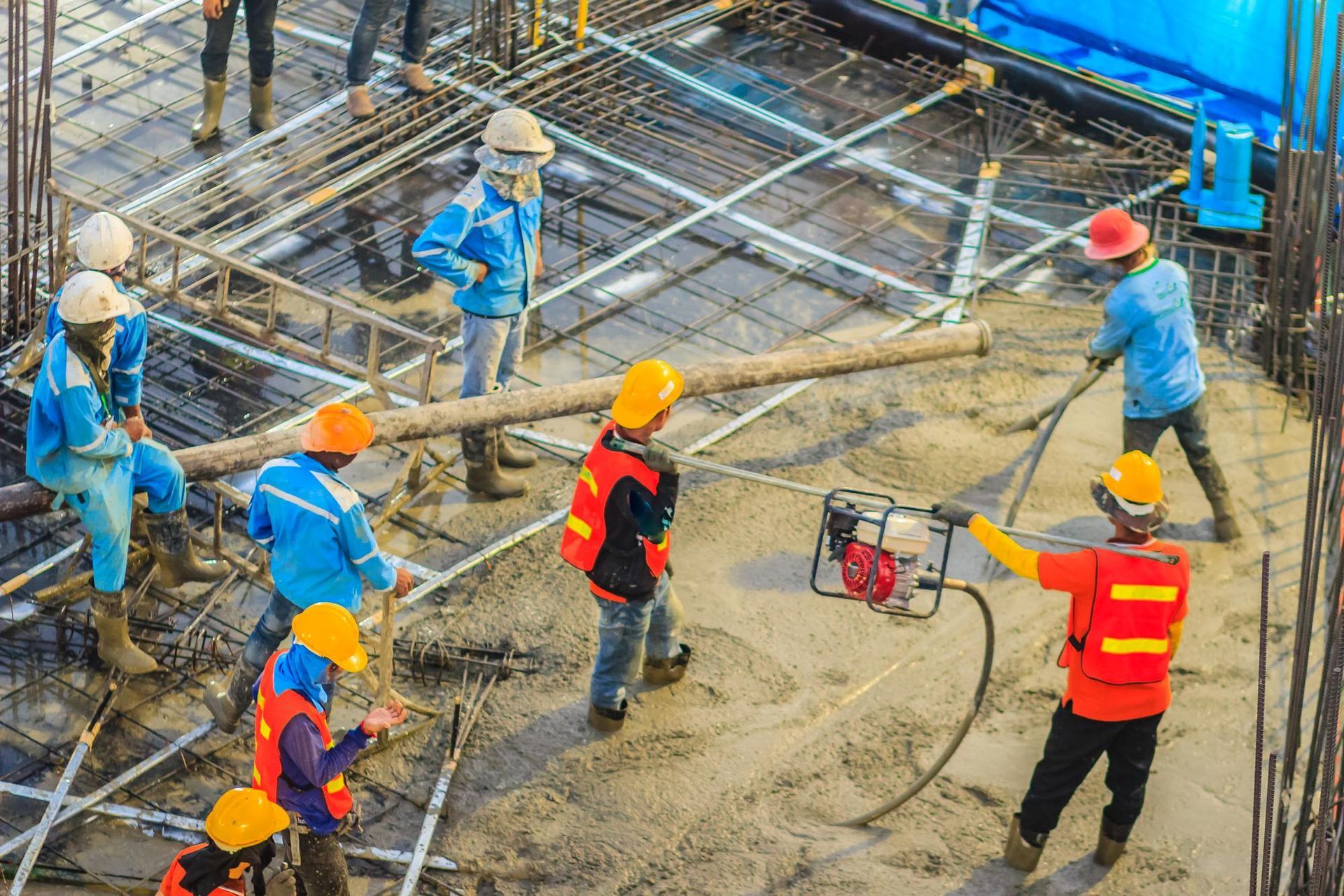 Construction workers pouring concrete on a building foundation; some use tools, wear hard hats and protective vests.