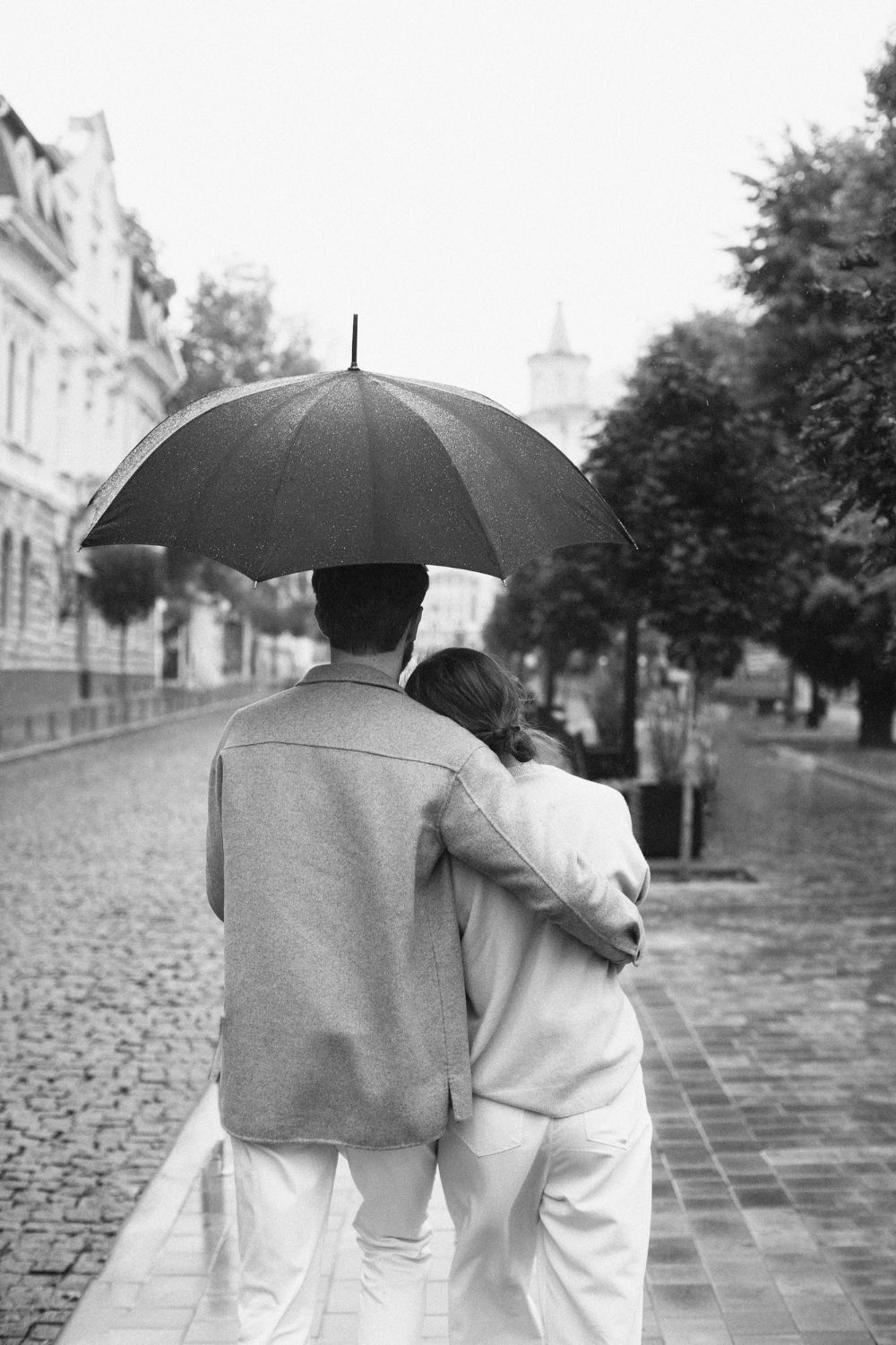 Couple walking in rain under umbrella, arm around each other on cobblestone street.