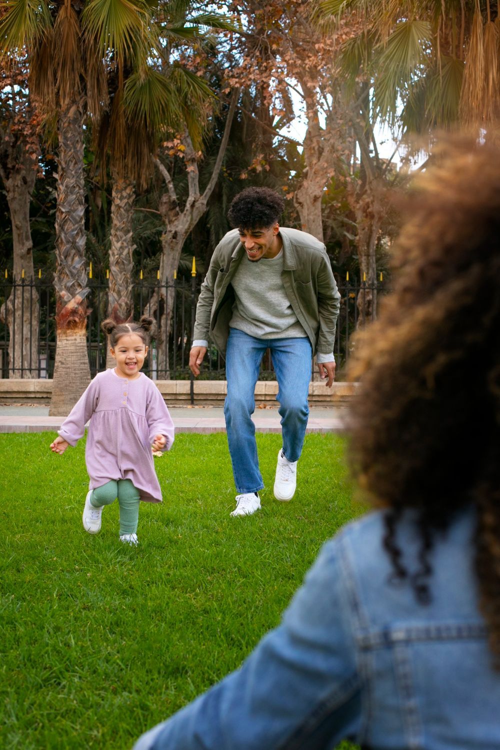 Child running towards smiling adult, another adult facing them on a grassy lawn with trees in background.