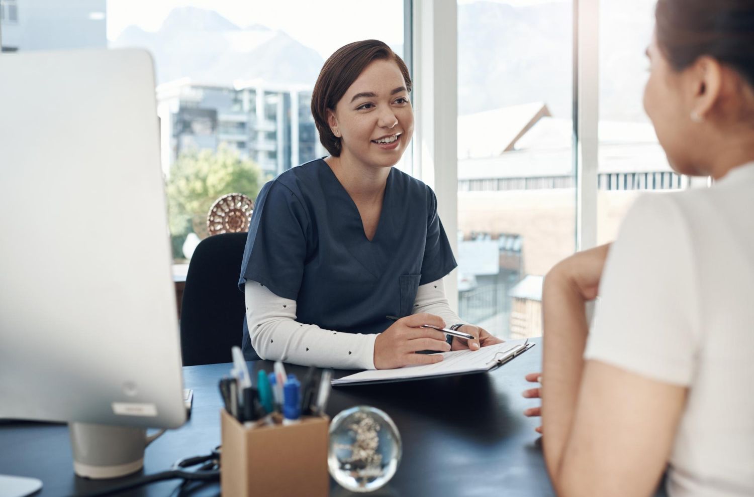 Woman in scrubs smiles and writes, speaking with a patient in an office.