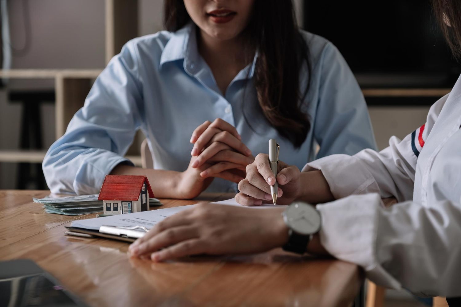 Two people at a table; one signing a document. A small house model sits beside the papers.
