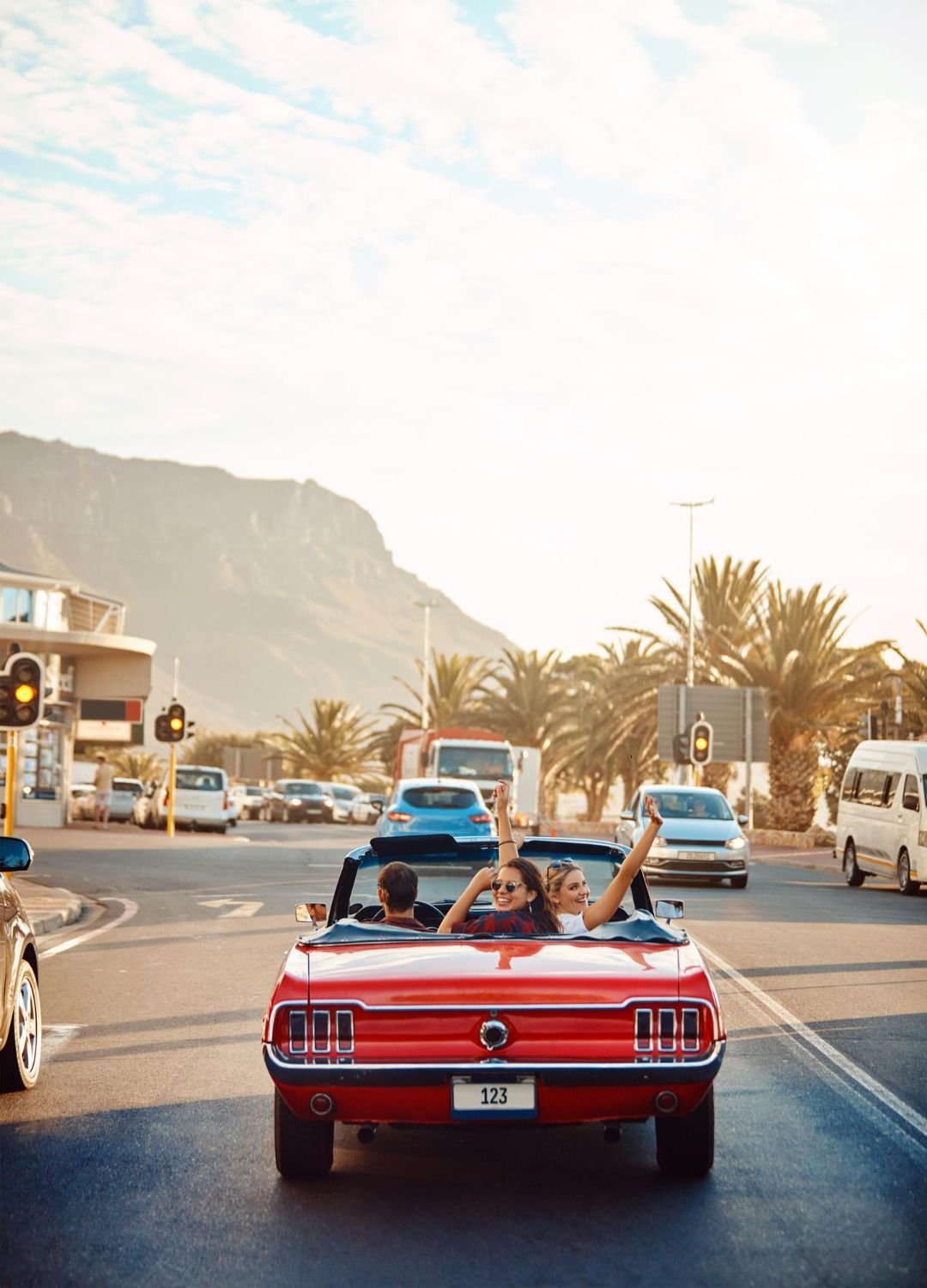 Red convertible car driving on a sunny street with passengers, mountains in background.