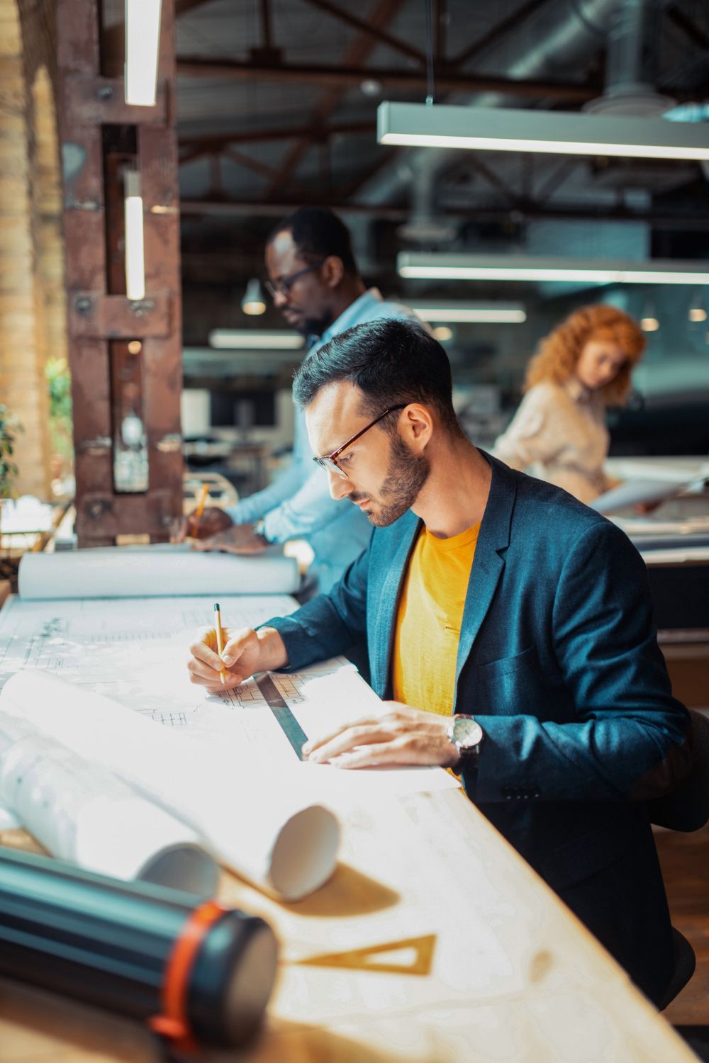 Man in blazer writing on blueprints; two colleagues in background. Drafting office setting.