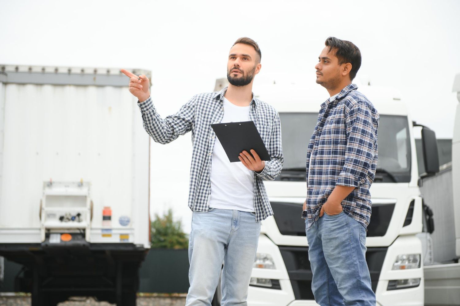 Two men near a semi-truck, one pointing, holding a clipboard, discussing details in a lot.