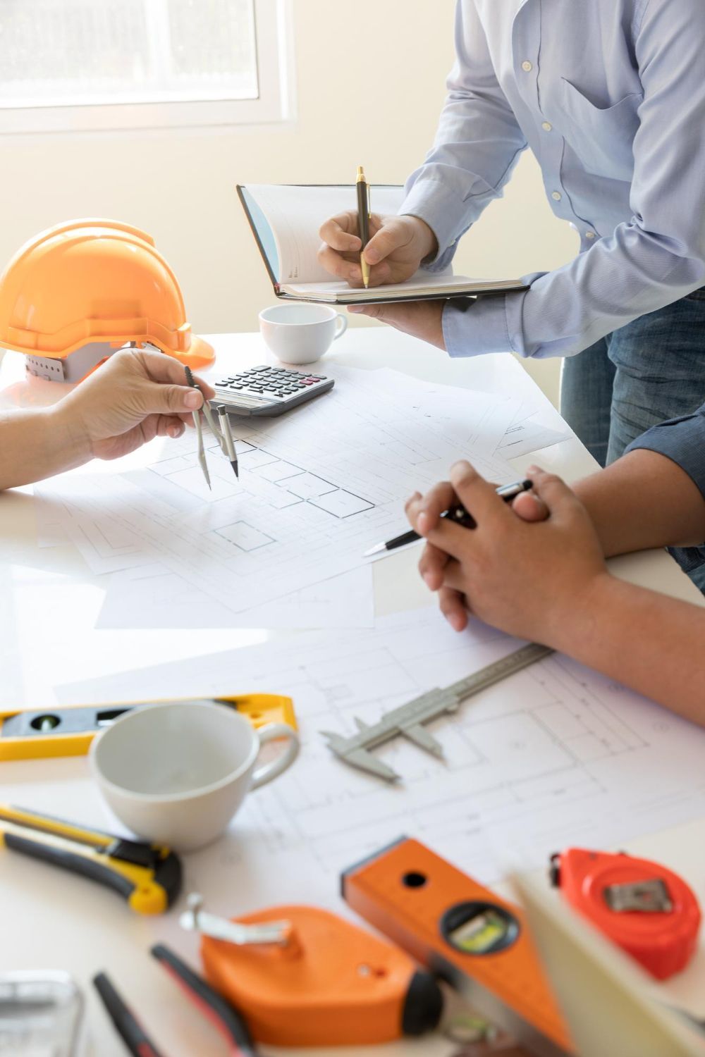 Architects reviewing blueprints at a table with tools and a hard hat. Someone is taking notes.