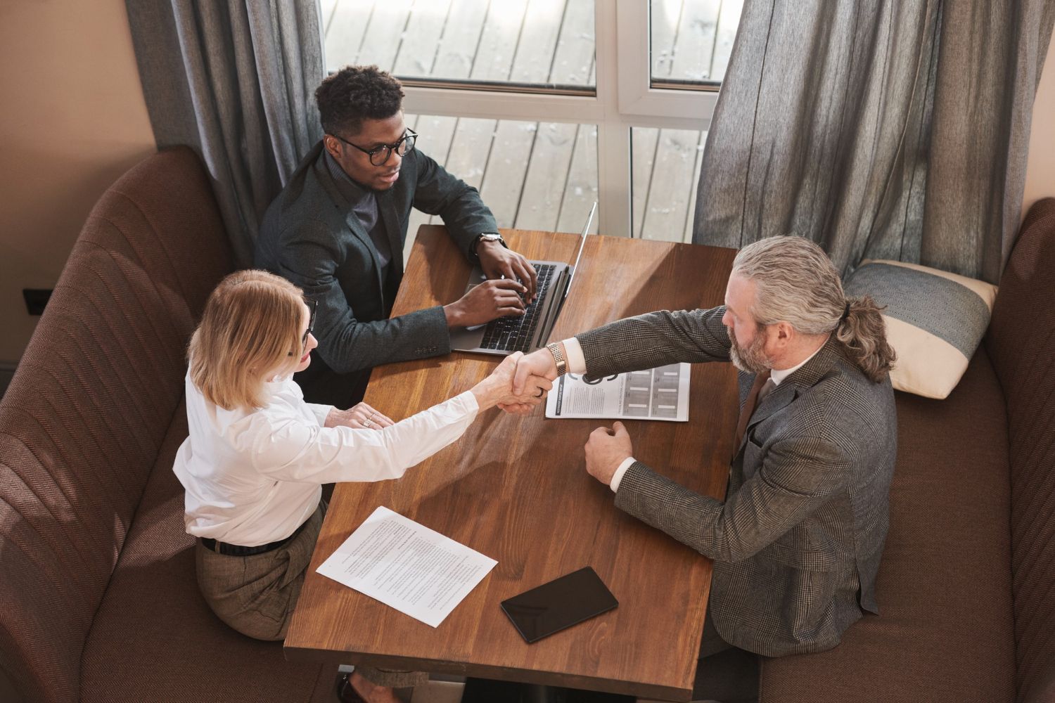 Three people at a table in a booth, one shaking hands with a woman, another typing on a laptop.