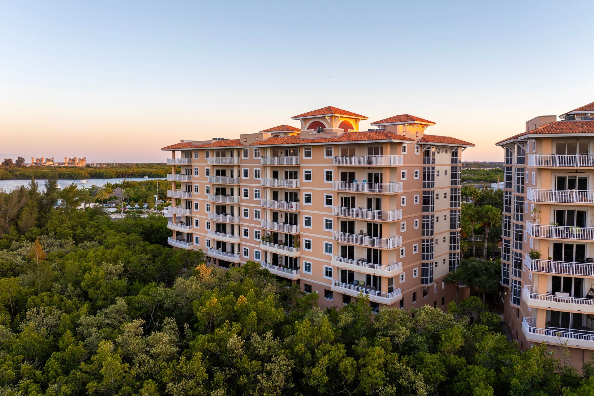 Multi-story beige condo building among green trees, overlooking water at sunset.