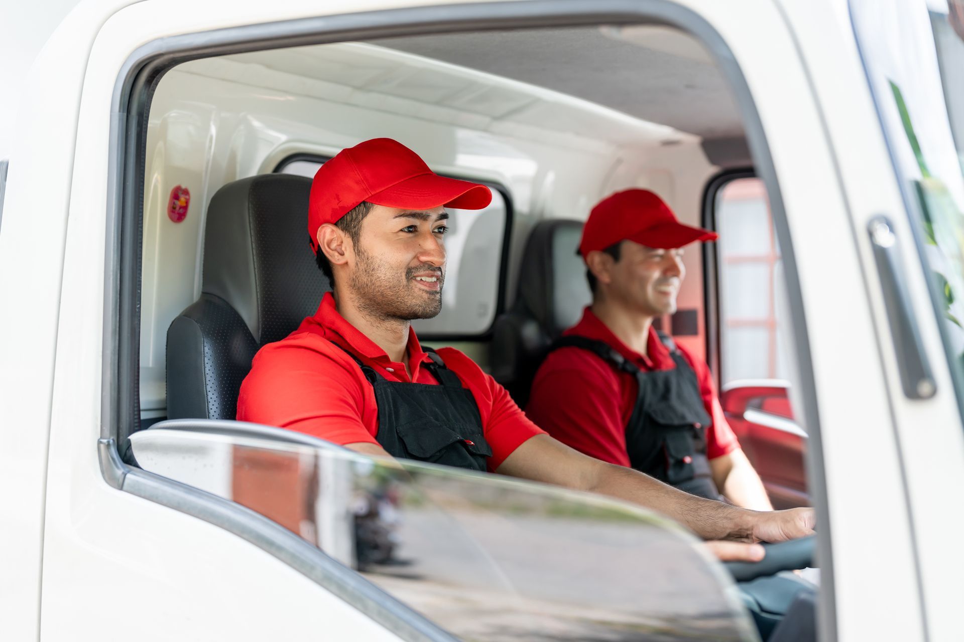 Two delivery drivers in a white truck, both wearing red caps and shirts.