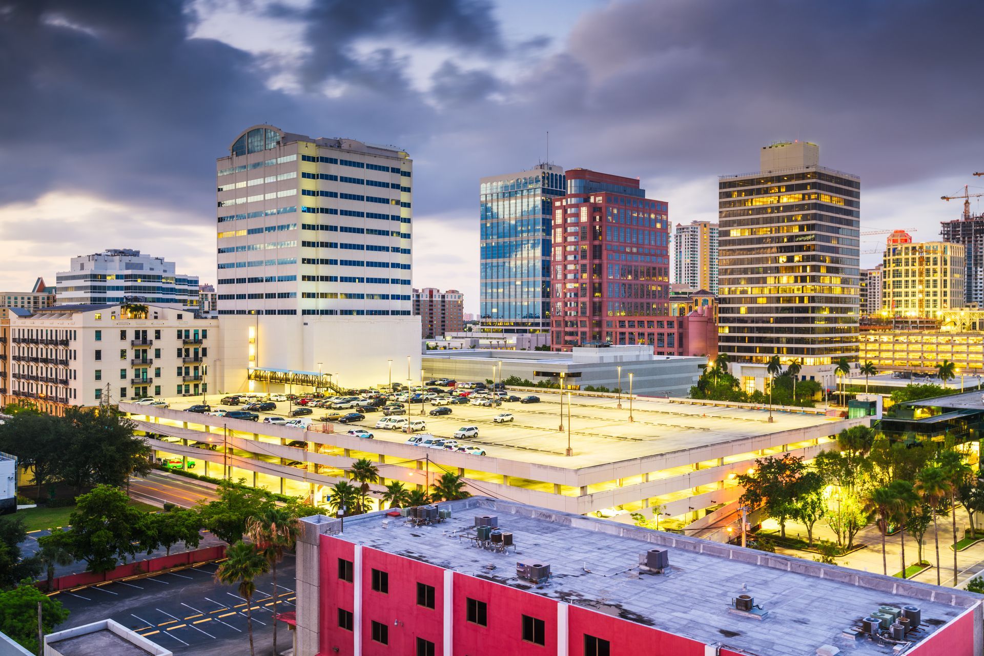 Skyline of downtown buildings at dusk. Includes parking garage, palm trees, and illuminated windows.