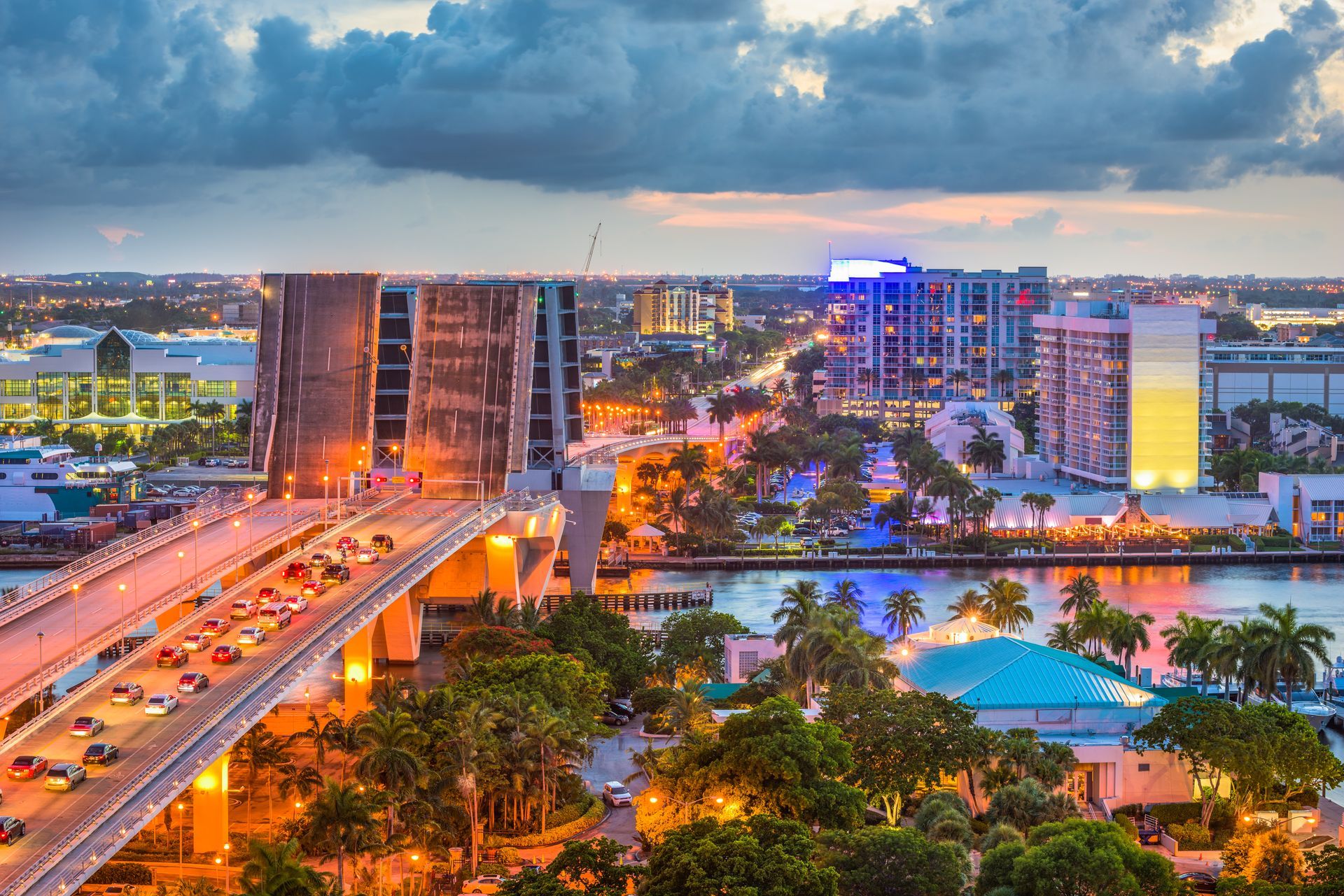 Cityscape at dusk, bridge with traffic, waterfront buildings, and colorful lights.