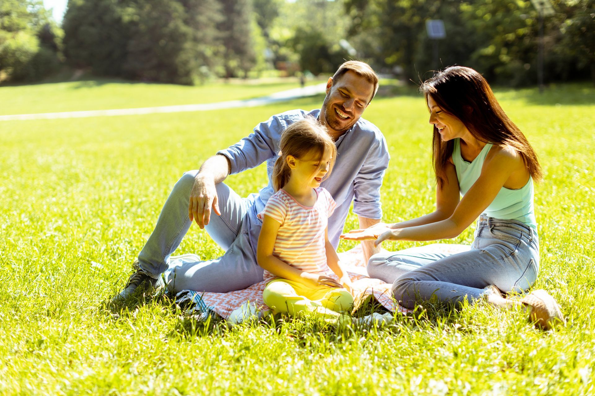 Family of three on a picnic blanket in a sunny park. They are laughing and smiling.