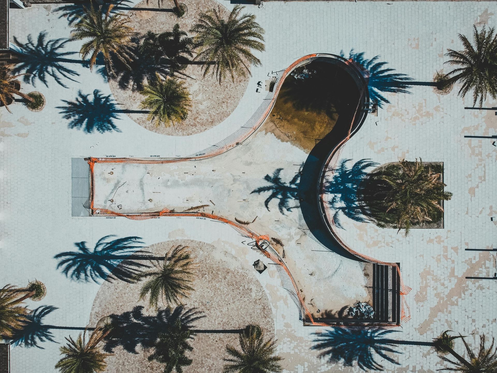 Aerial view of a skatepark with curved concrete ramps and palm tree shadows.
