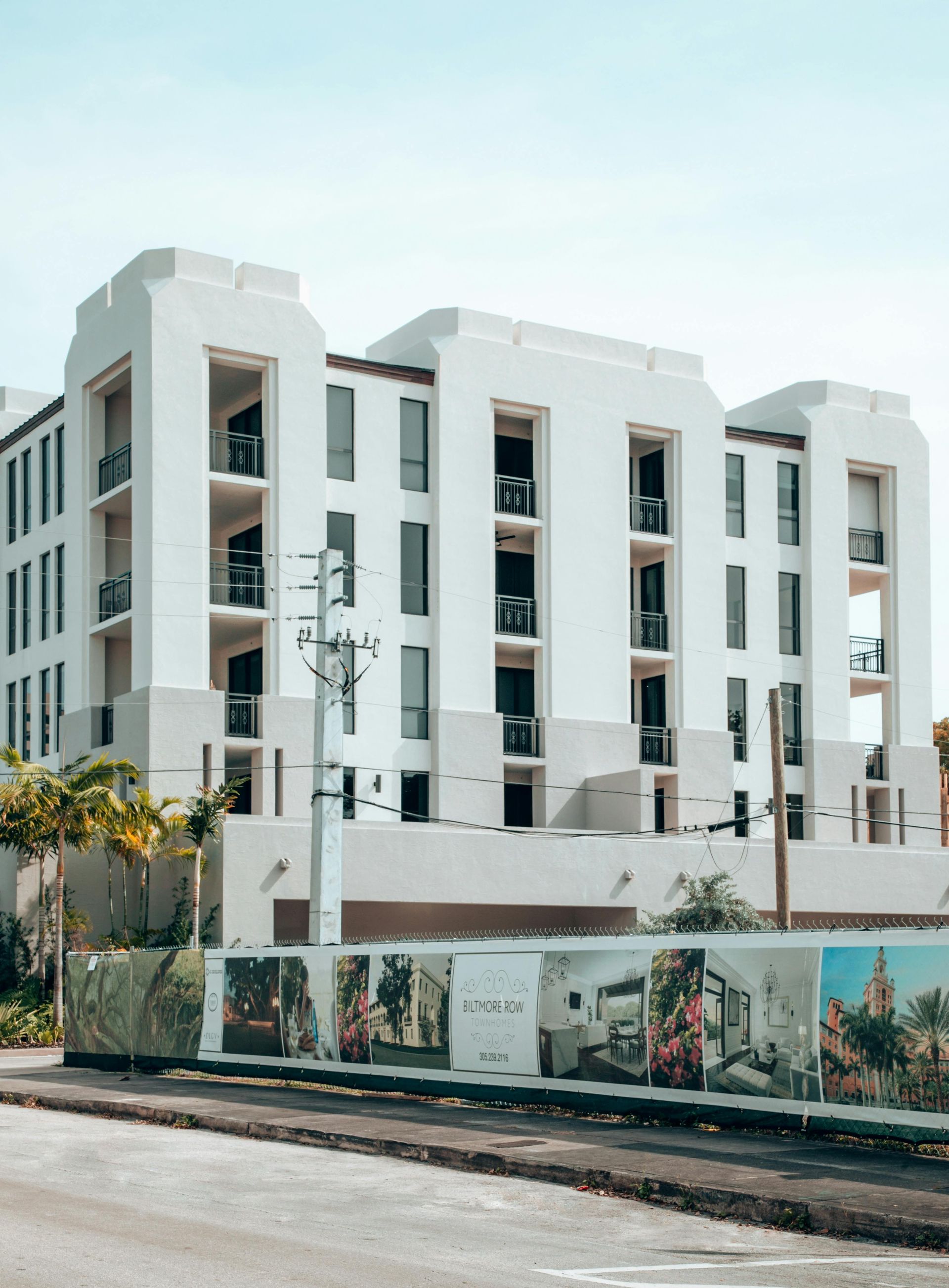 White apartment building with balconies, construction fence in front, blue sky.