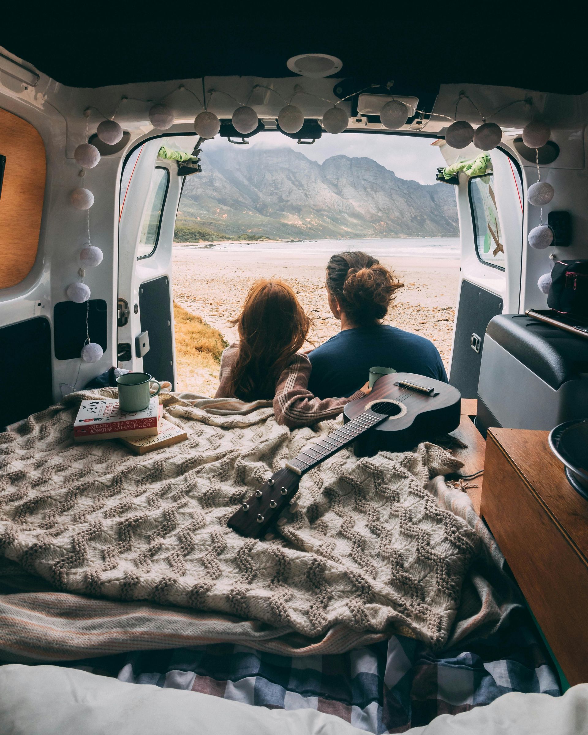 Couple in a camper van, gazing at a beach view with mountains. Guitar and blanket are on the bed.