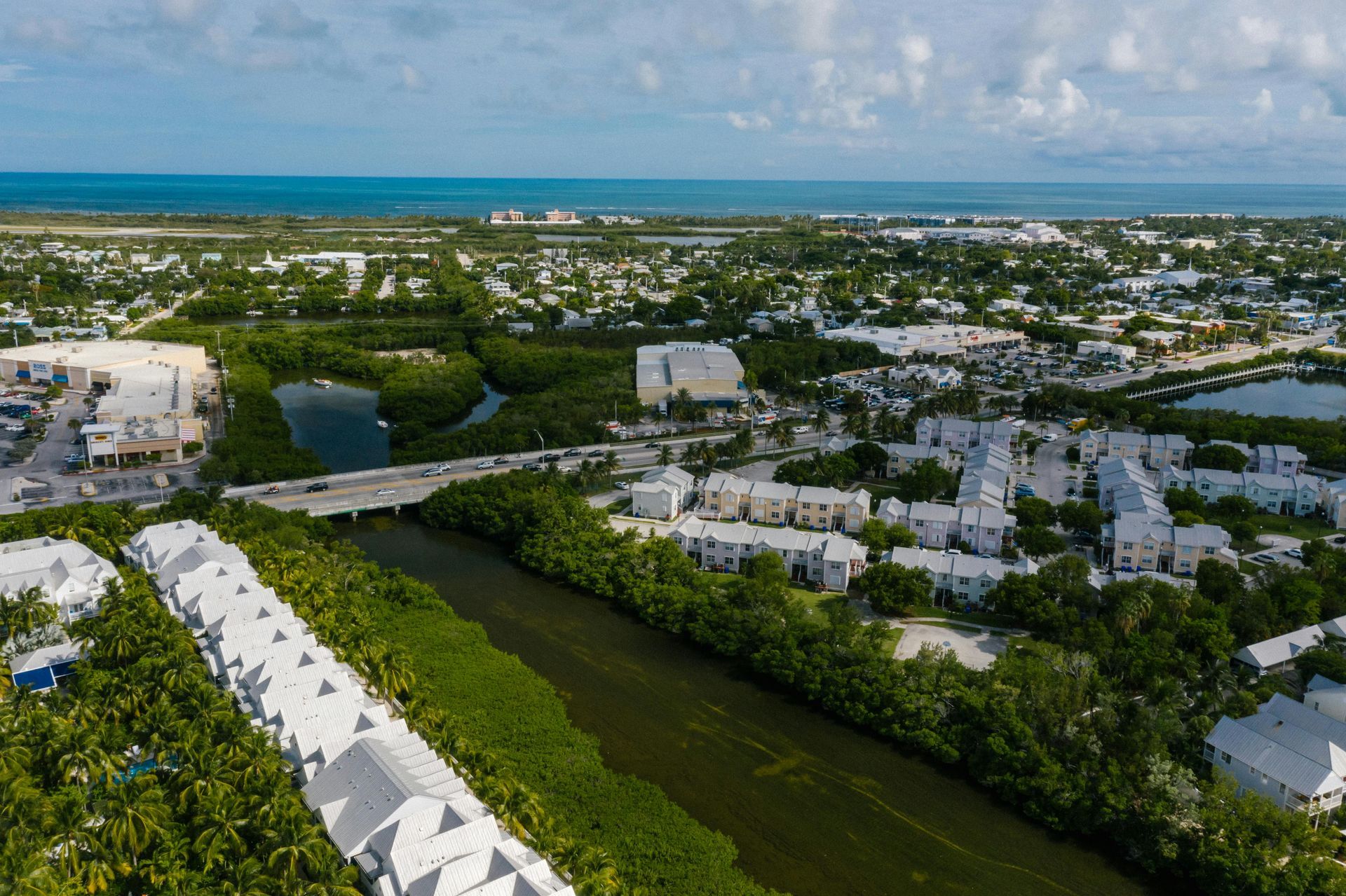 Aerial view of a coastal town with white buildings, lush green mangroves, a bridge, and the blue ocean.