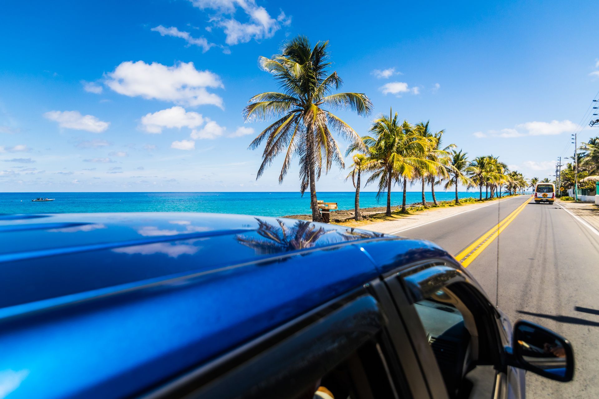 Blue car driving along a coastal road lined with palm trees under a bright blue sky.