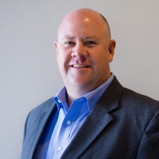 Man in a blue shirt and gray blazer smiles, against a gray background.