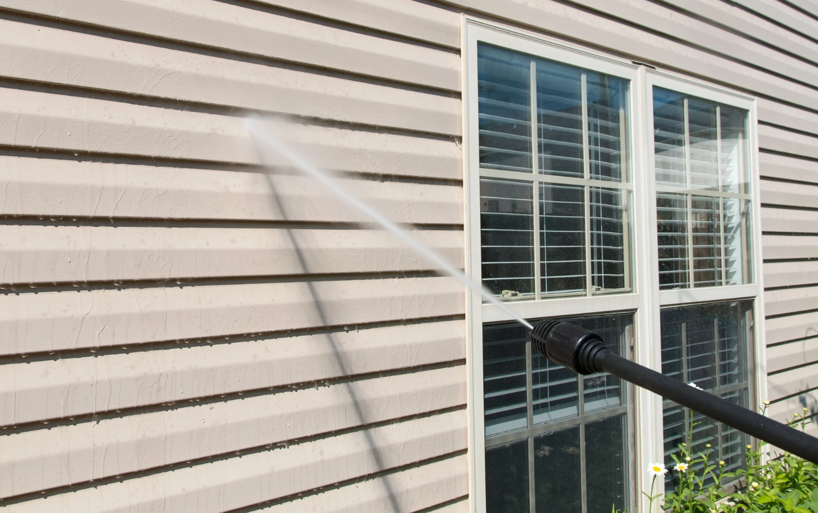 Power washer spraying beige vinyl siding near a window.