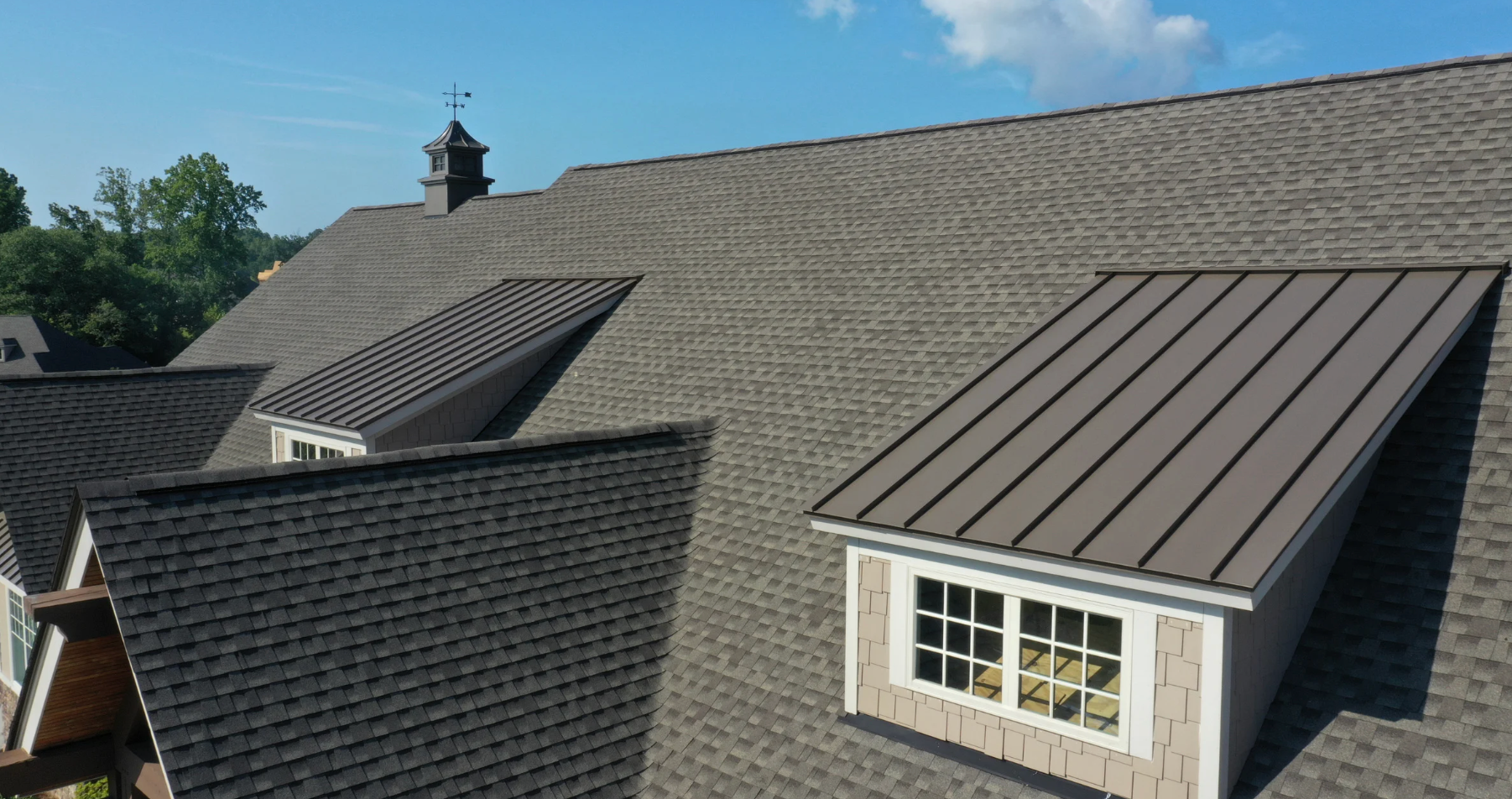 Gray asphalt shingle roof with two brown metal accent roofs and a dormer against a blue sky.