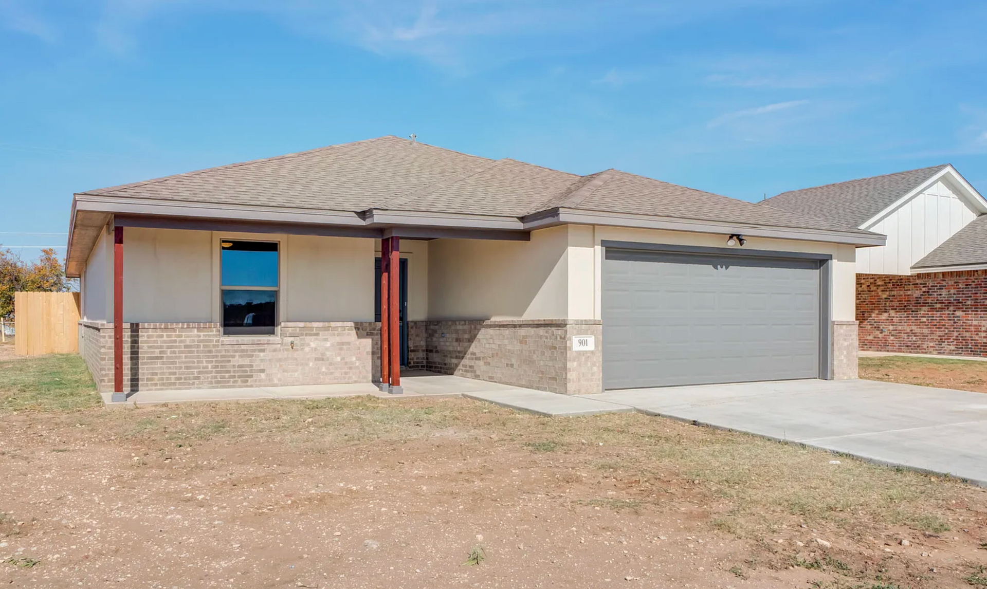 Tan, one-story house with gray garage door, brown brick and supports, on a brown, dry lot under a blue sky.