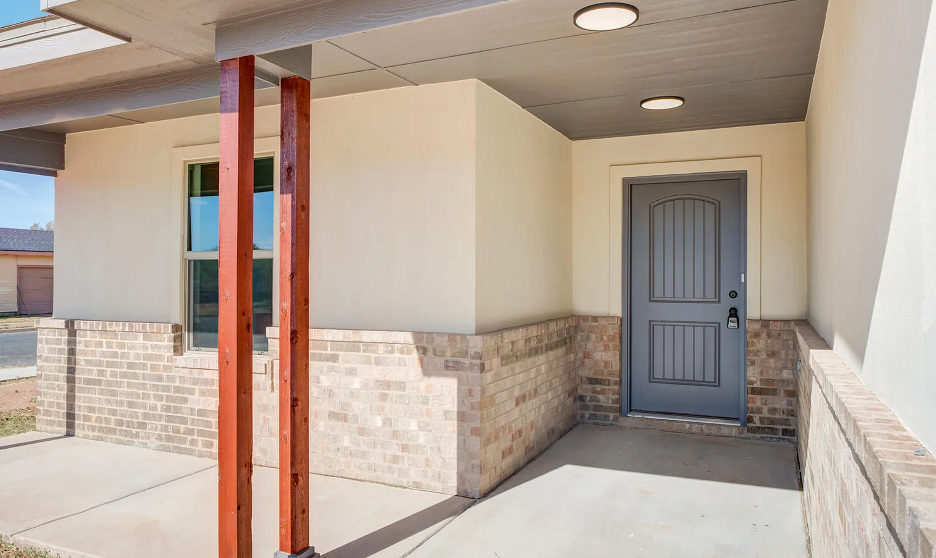 Covered entrance of a house with gray door, brick and stucco walls, concrete floor, and red support beams.