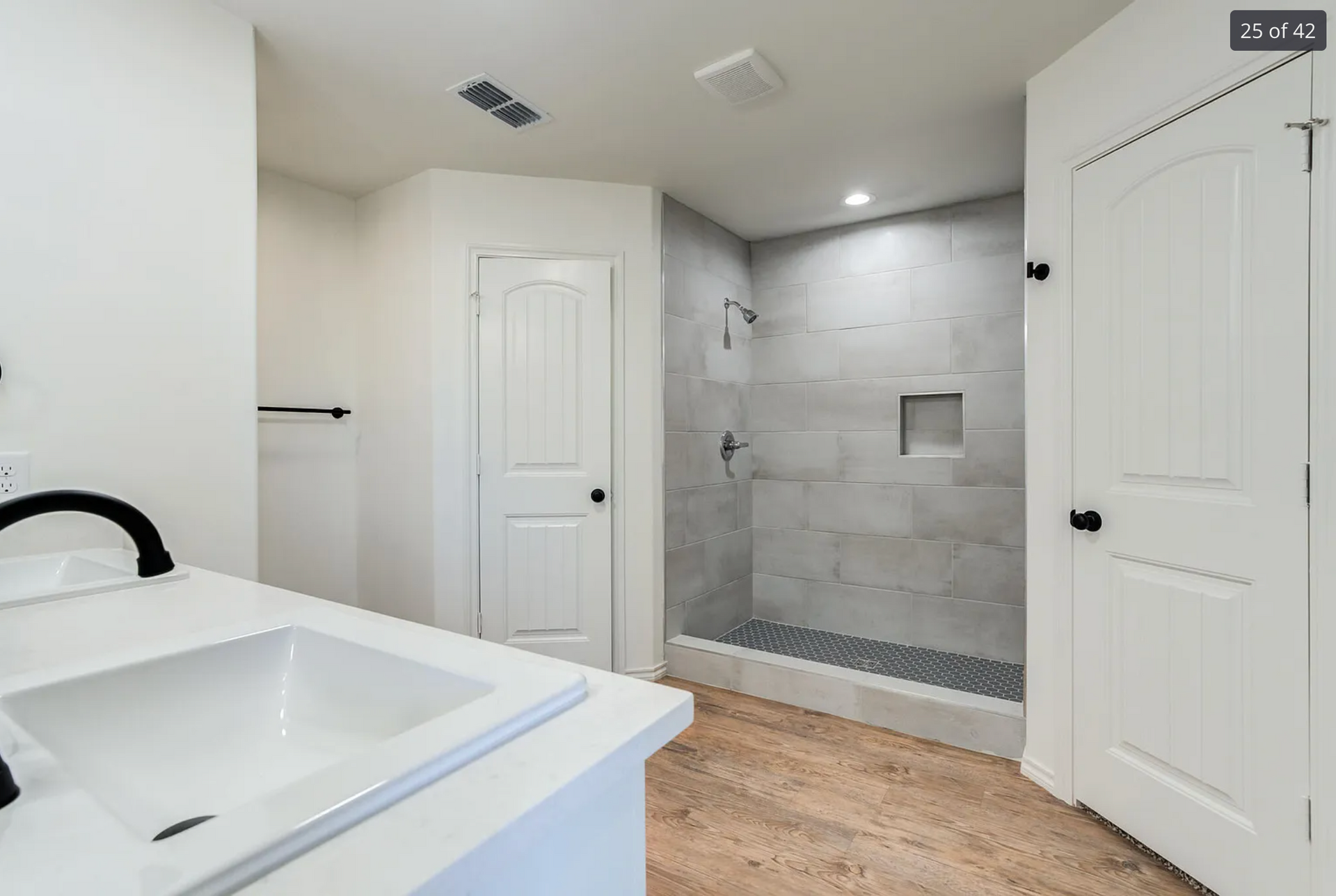 Bathroom with white vanity, shower, and closed doors; light wood floor, gray tiled shower wall.
