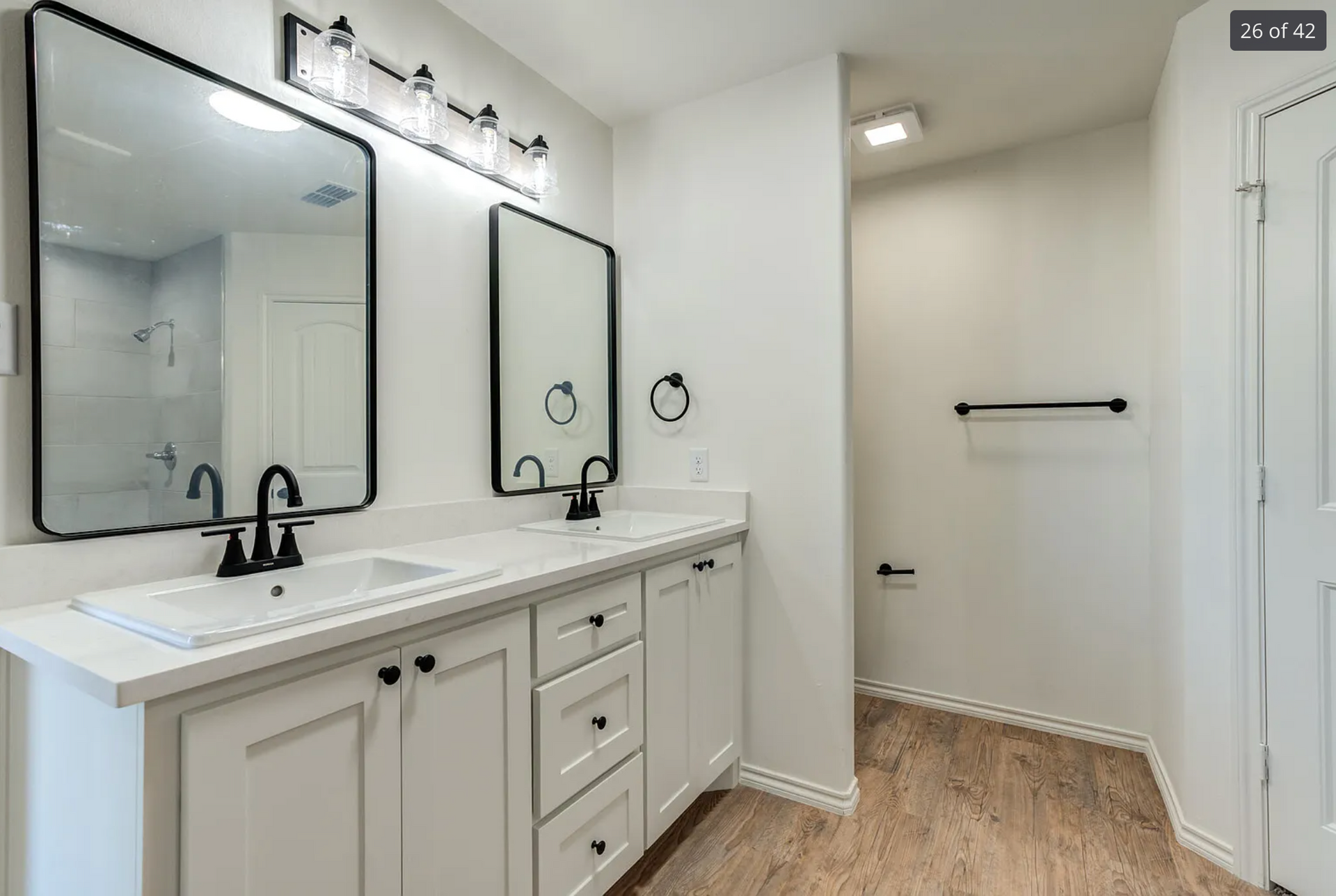 Bathroom with white cabinets, black fixtures, and two mirrors. Light wood floor and white walls.