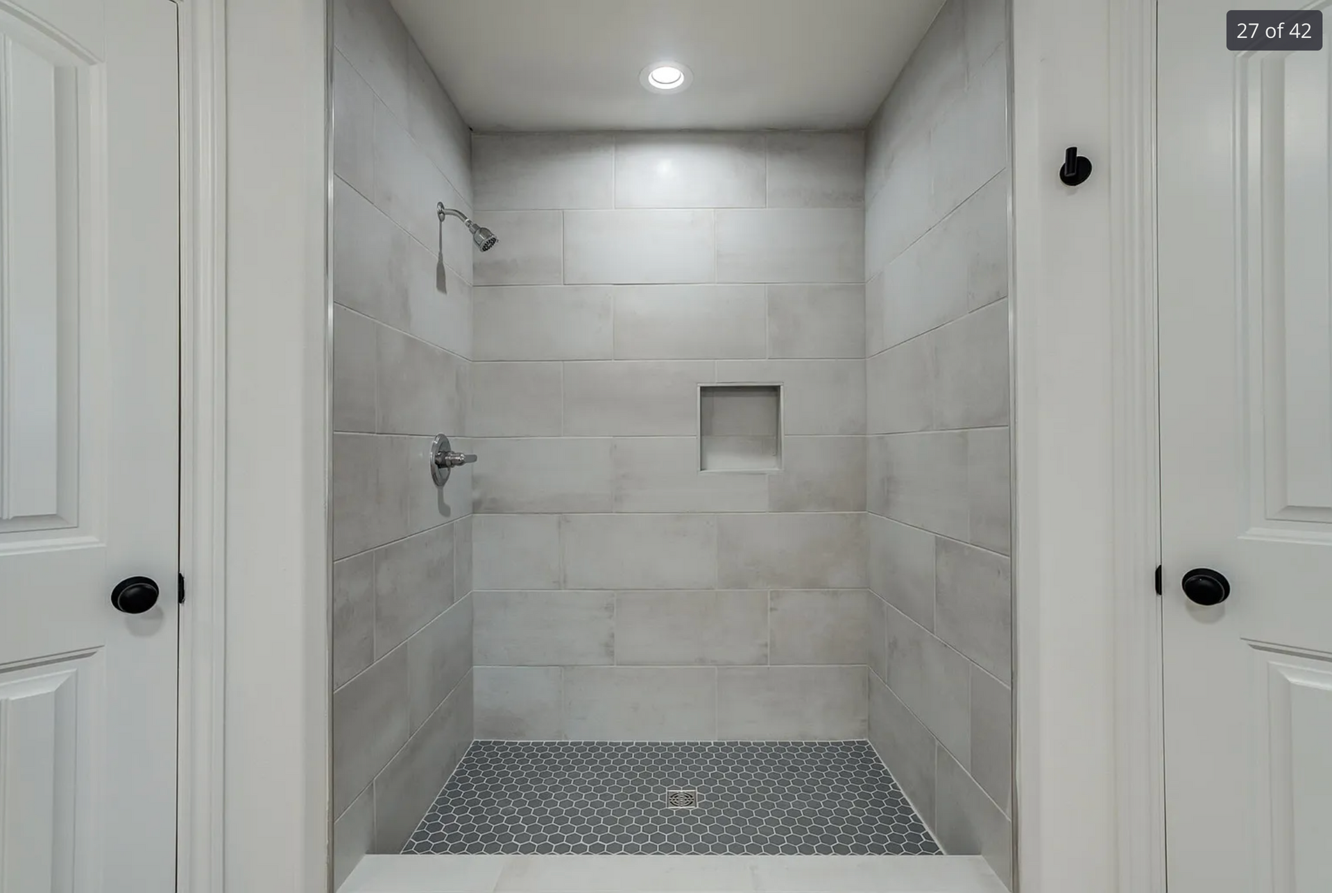 Gray-tiled shower with pebble floor, recessed shelf, and chrome fixtures between white doors.