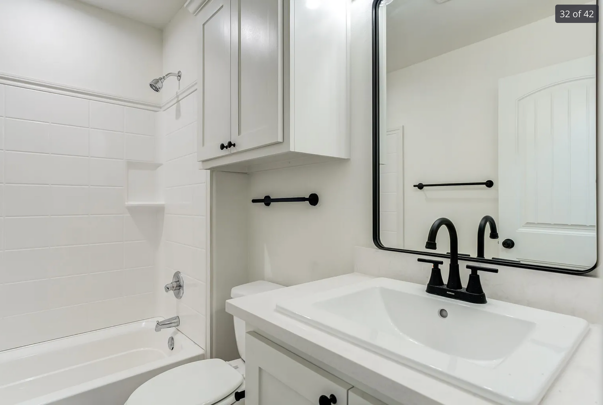 White bathroom with a sink, toilet, tub/shower, and cabinet. Black fixtures and mirror frame.
