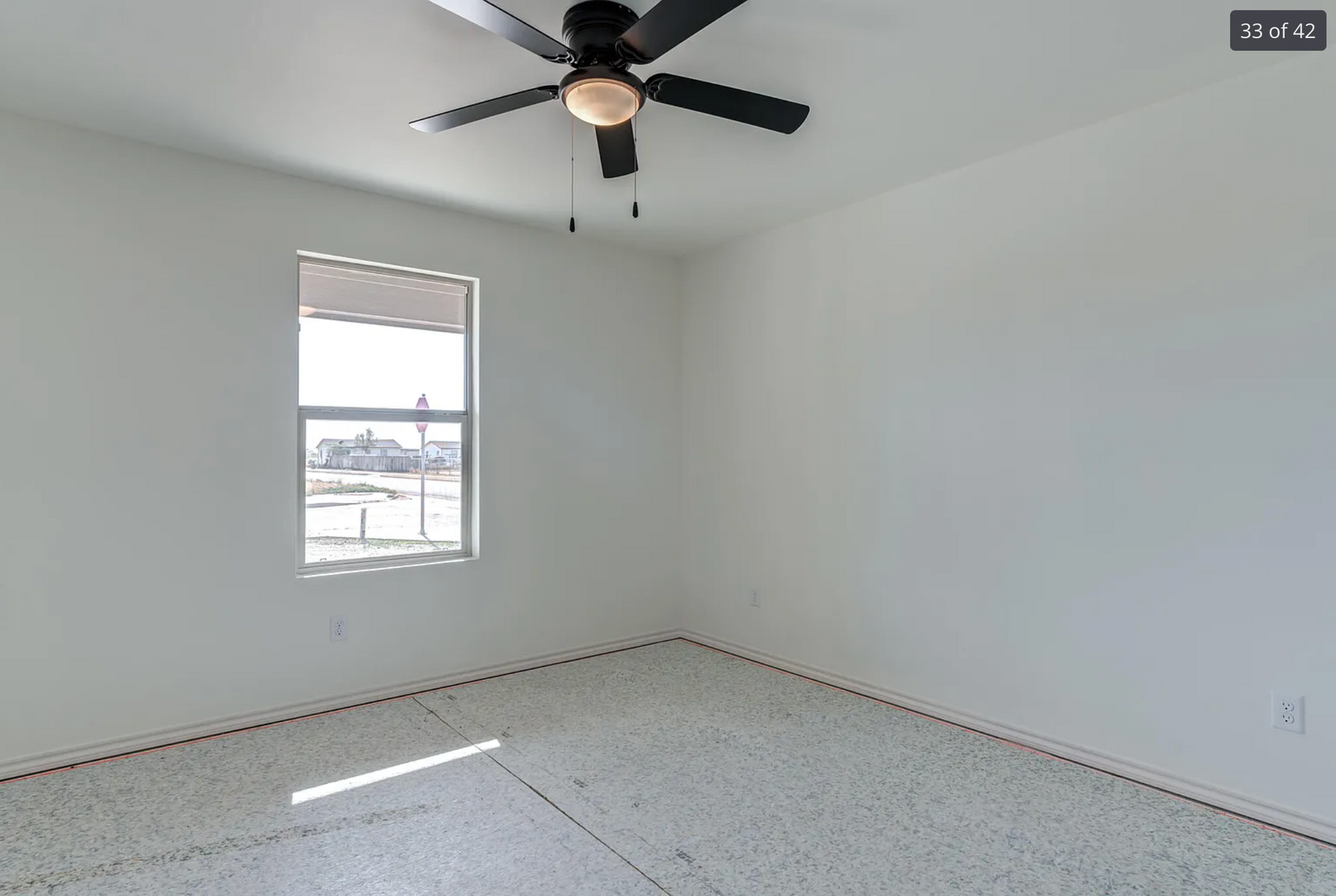 Empty bedroom with white walls, window, ceiling fan, and speckled flooring.