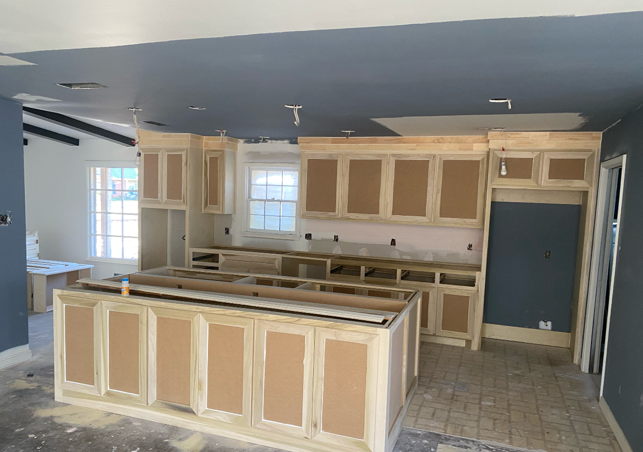 Kitchen under construction, cabinets installed, unfinished walls and ceiling, blue-gray paint, window.
